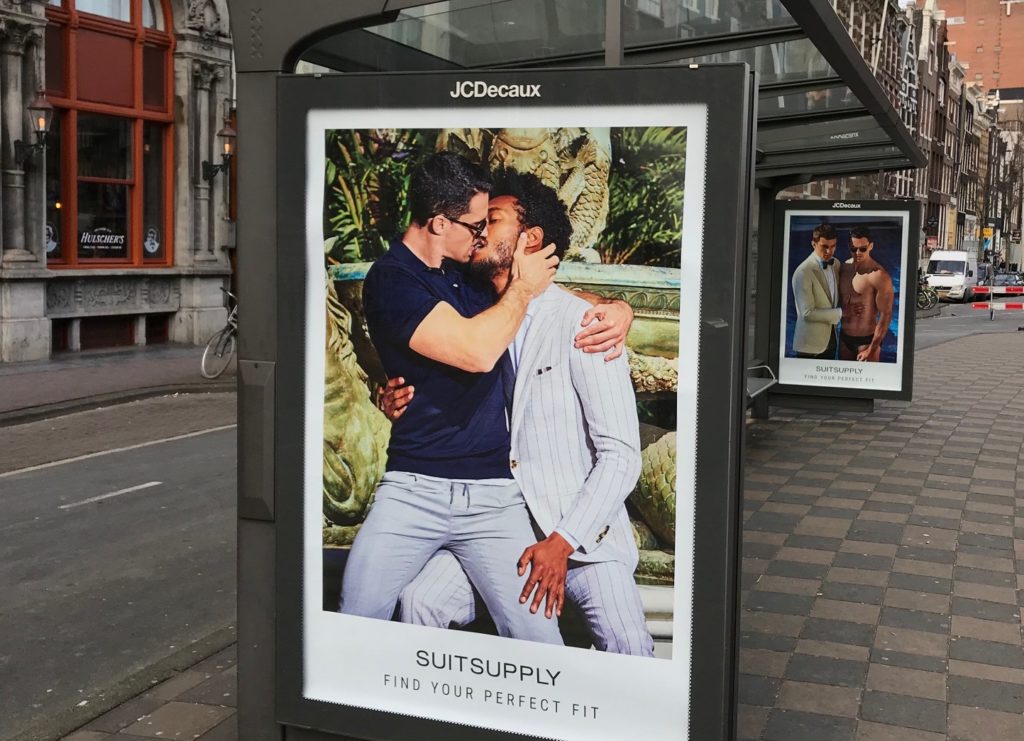 Een foto van de reclame van Suitsupply in een bushokje. Op de reclame zijn twee mannen zoenend te zien.
