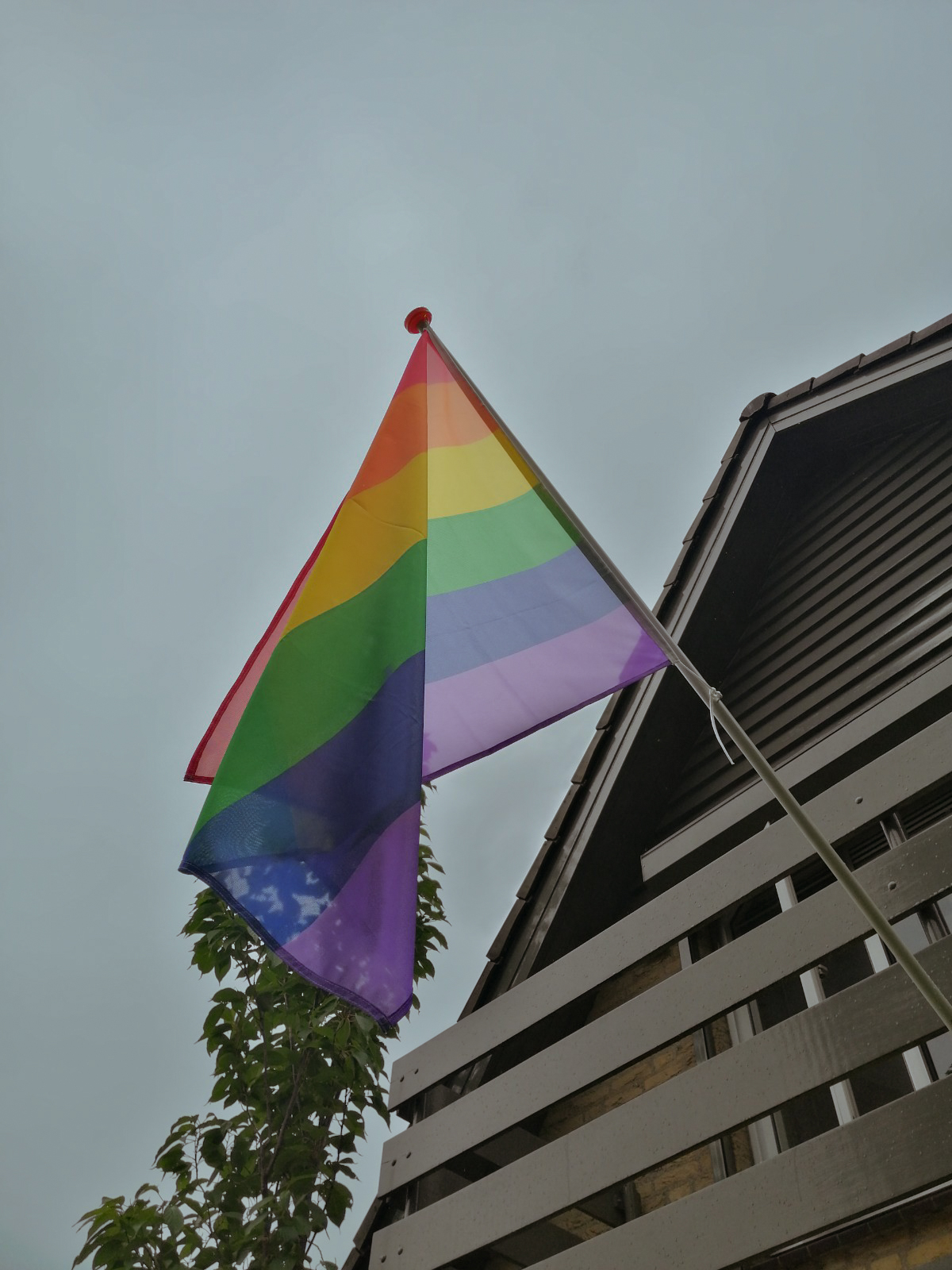 Er hangt een grote regenboogvlag aan een huis aan het balkon op een vlaggenstok. Deze is van onderen gefotografeerd.